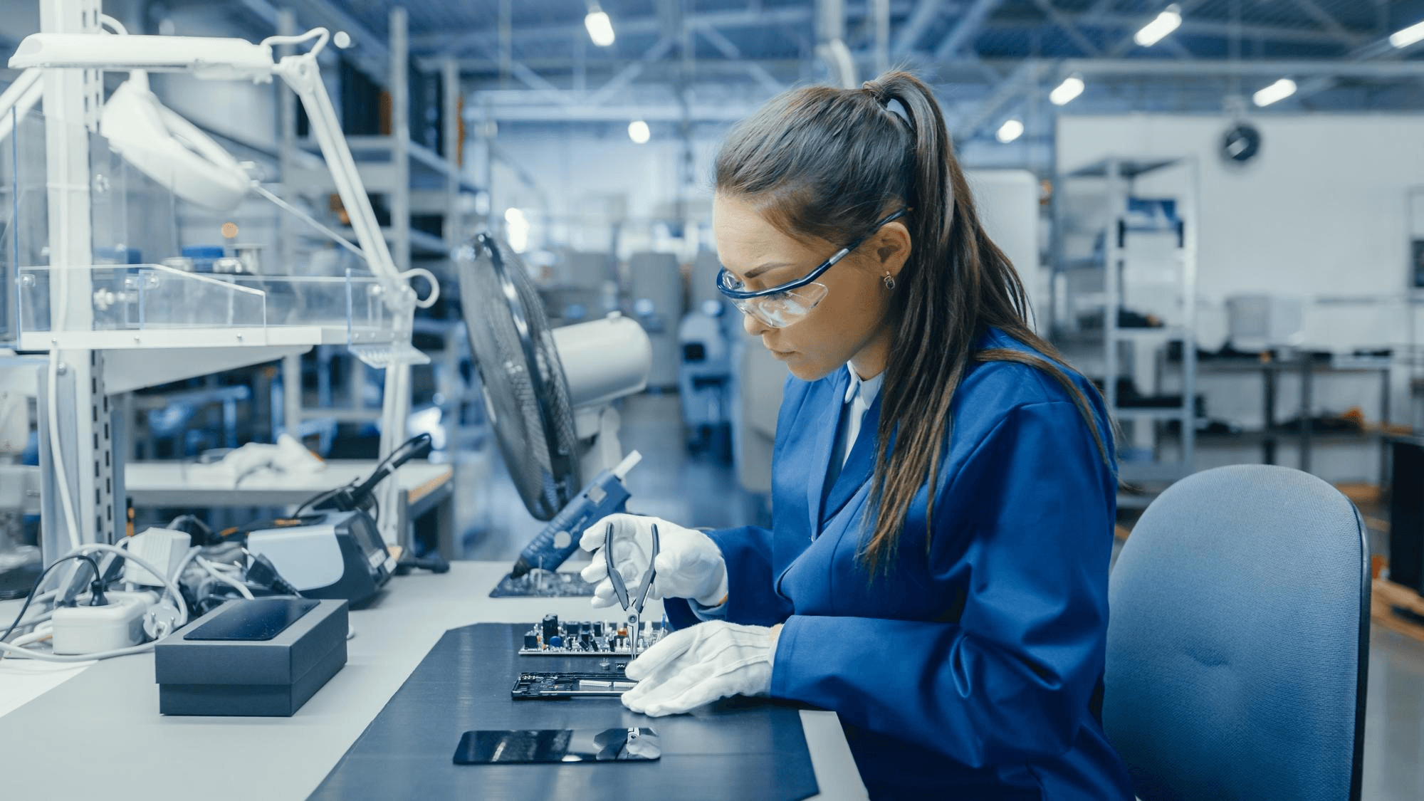Technician in safety glasses inspecting a circuit board under a microscope